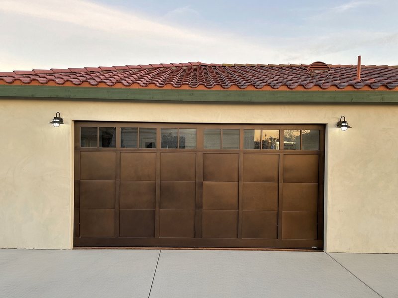 Front view of a bronze carriage-style garage door with a row of windows across the top, lit by barn lights at dusk