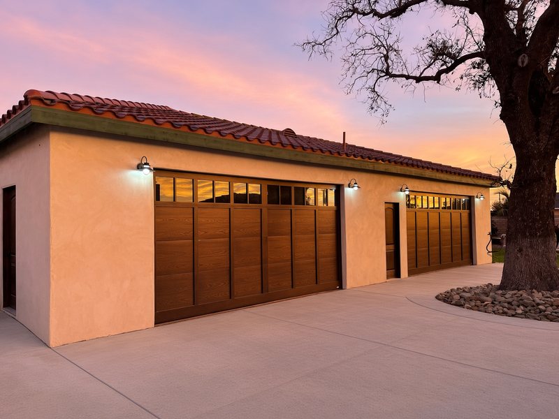 Bronze carriage-style garage doors with window tops on a stucco garage with a red tile roof at sunset